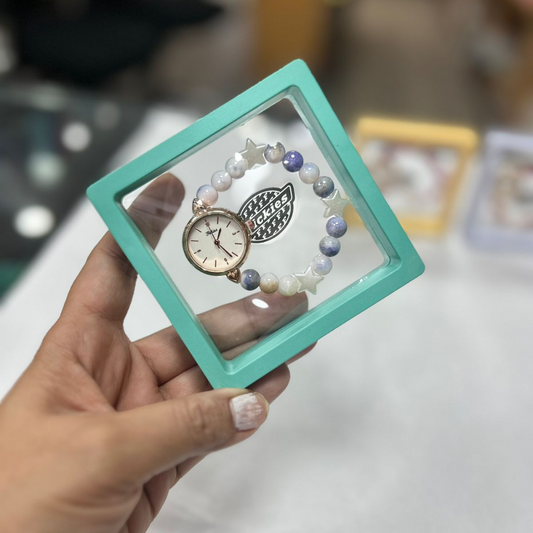 A hand holds a mint-green square display frame with a delicate circular, gear-like ornament inside, against a blurred indoor background; the scene feels crafty.
